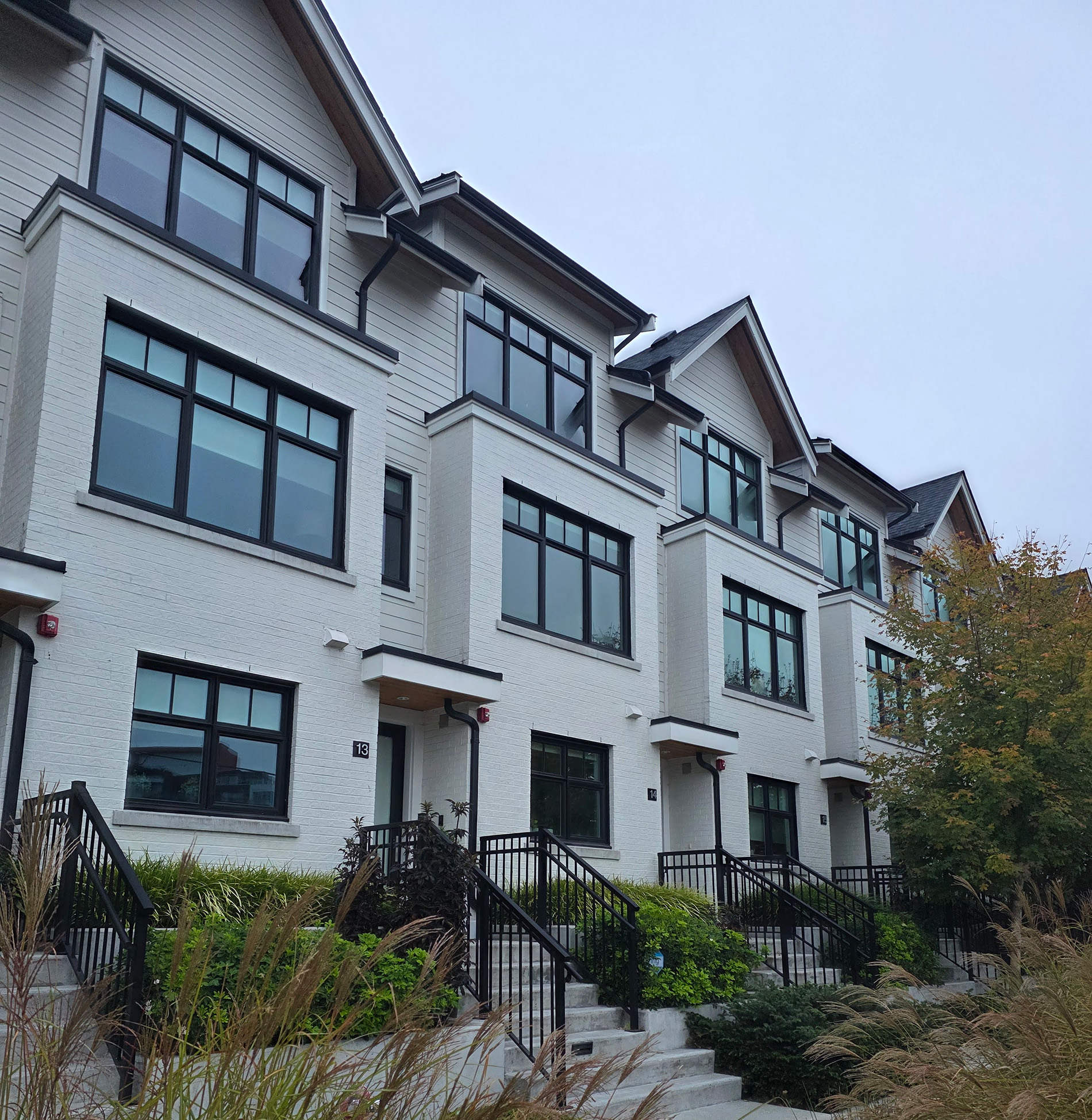A row of modern, multi-story townhouses with large windows, white brick exteriors, and black railings lining the steps up to each front door. Some greenery and ornamental grasses are visible in the foreground.