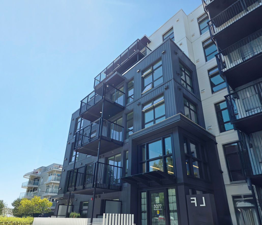 Modern apartment building with black metal balconies and large windows, photographed on a clear, sunny day. Some greenery and a white fence are visible in the foreground.