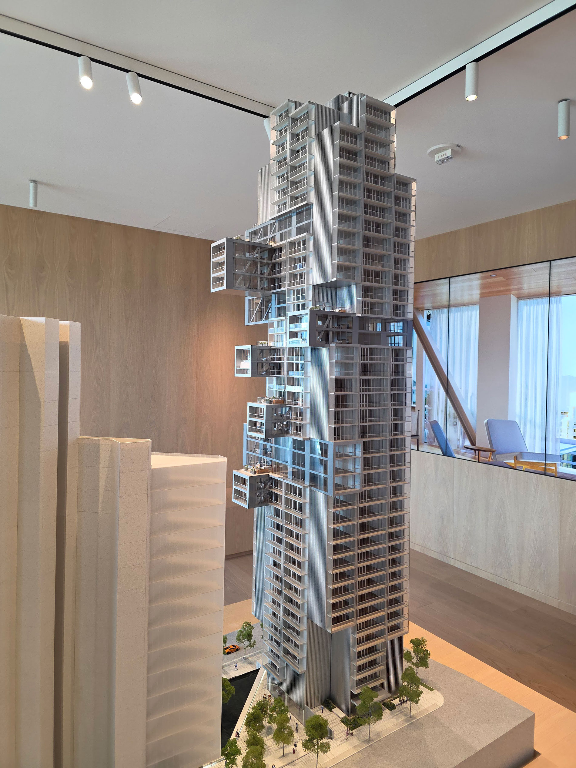 A detailed architectural model of a modern high-rise building with several protruding glass-box structures, surrounded by trees and other building models, displayed in a well-lit room with wood-panel walls.
