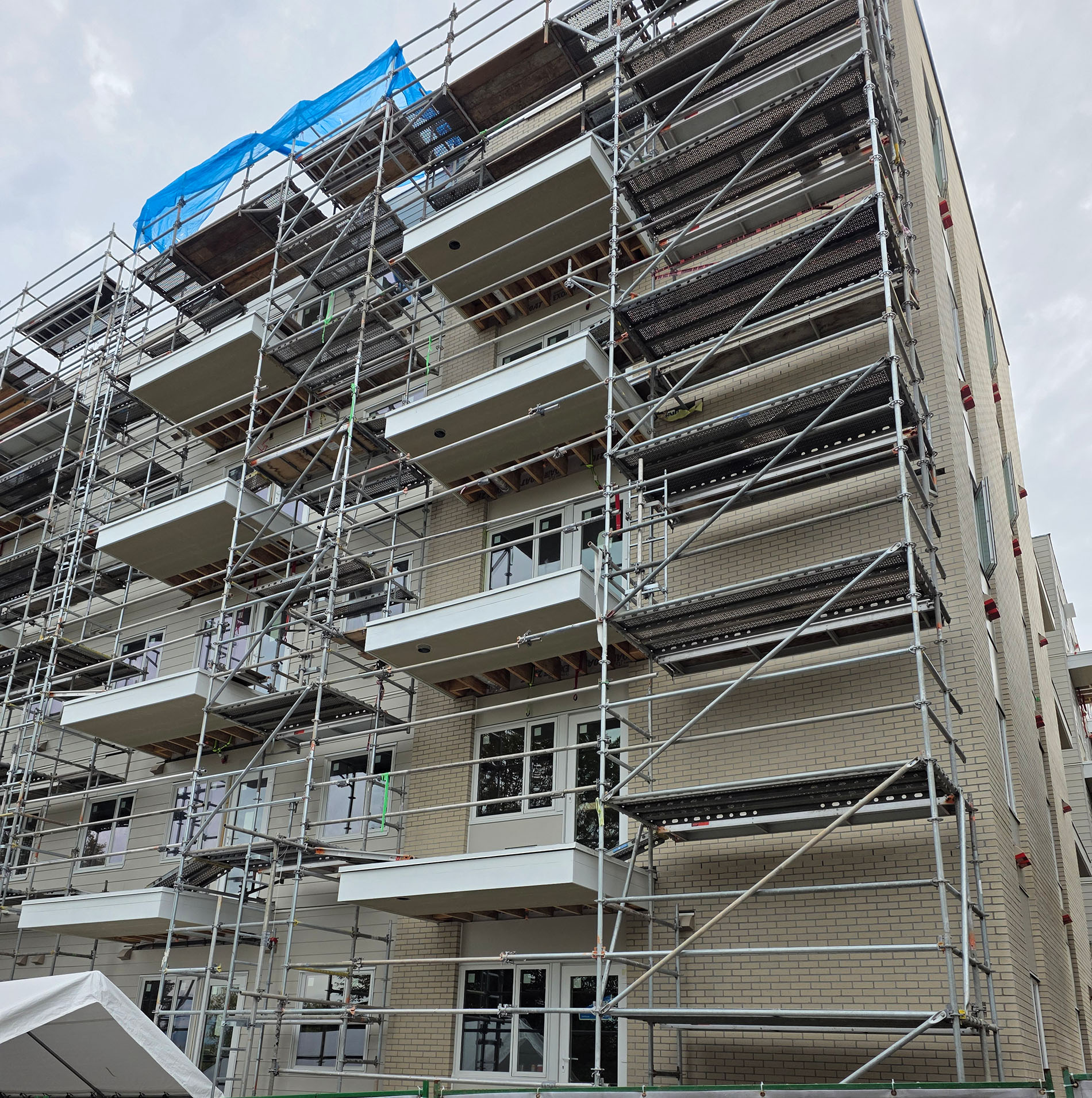 A multi-story apartment building under construction, surrounded by extensive scaffolding. Several white balconies are visible, and the facade is partially finished. A blue tarp covers part of the roof area.