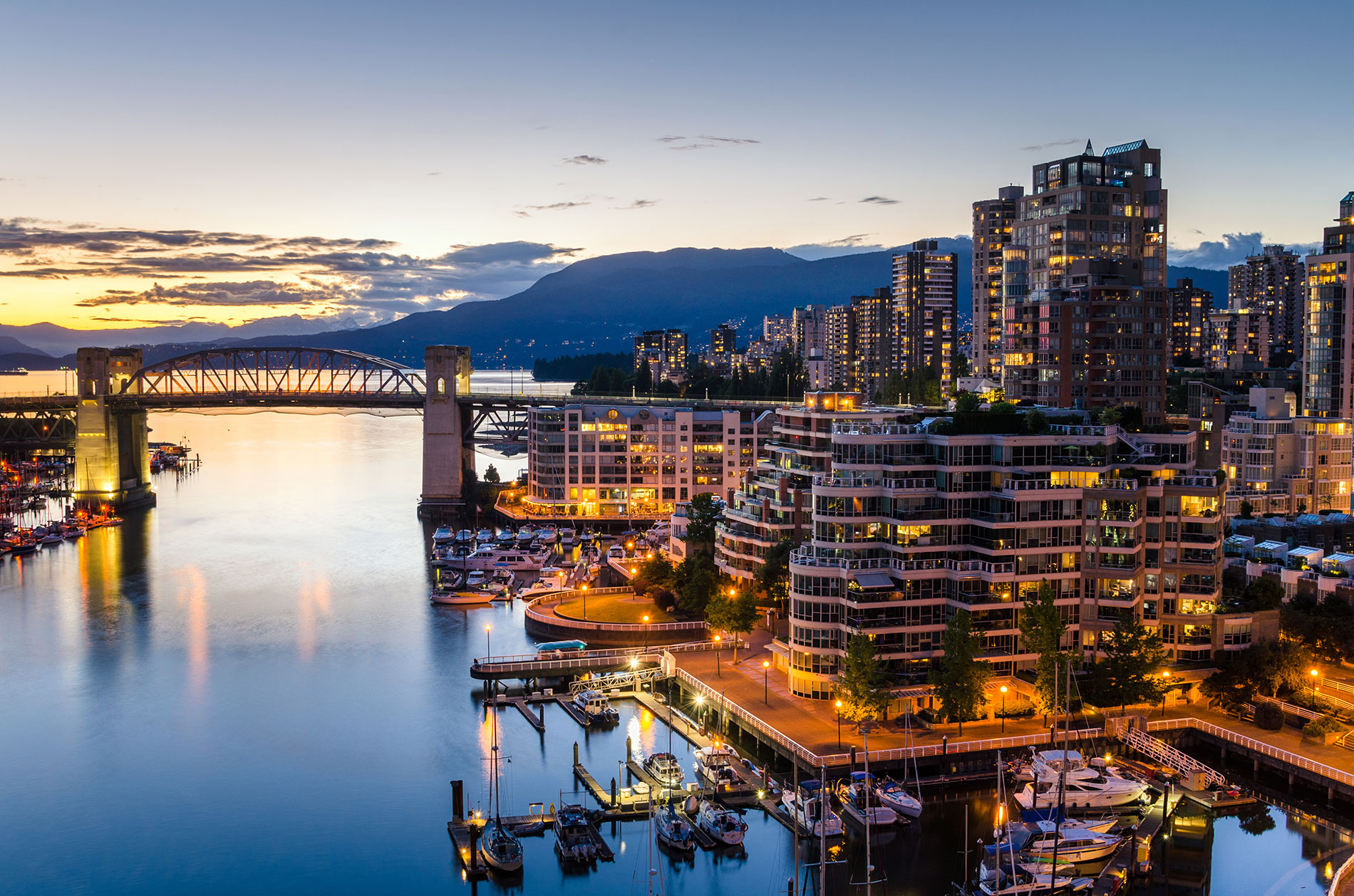 City skyline at dusk with high-rise buildings, a marina with boats, and a bridge spanning calm water; mountains are visible in the background under a blue and orange sky.